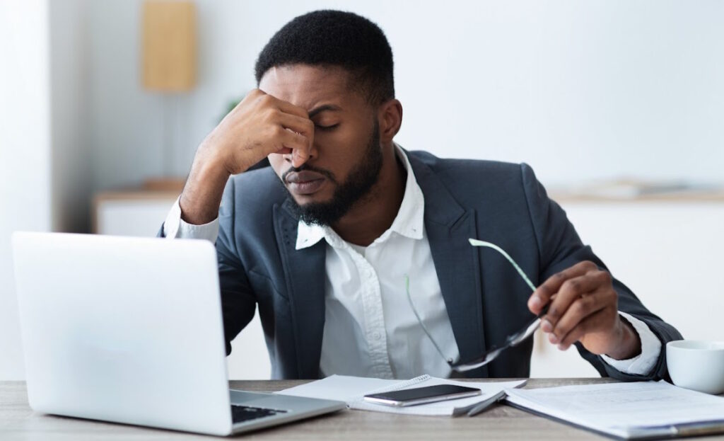 Man holds his glasses in one hand while rubbing his eyes and looking at a laptop. The man looks exhausted.