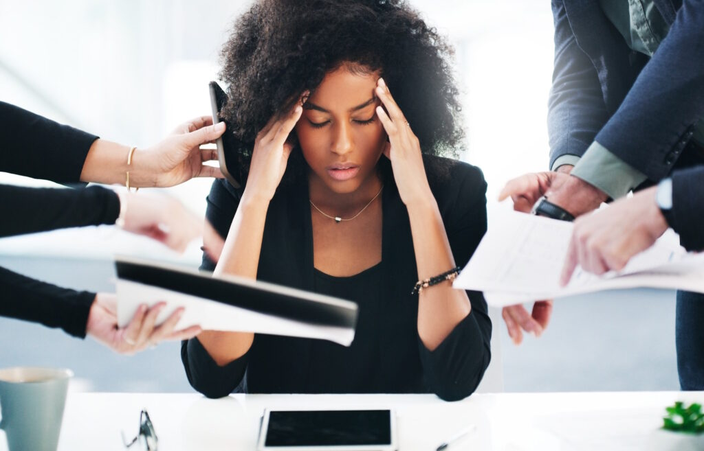 Black woman with her hands against her head with office documents being shoved in her face by multiple people.