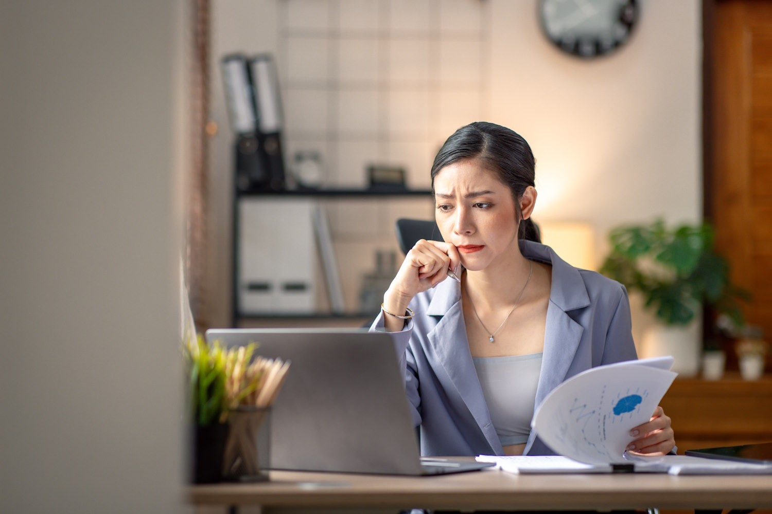 A woman is sitting at a desk looking at a laptop while flipping through a packet of paper. The woman looks serious and stressed.