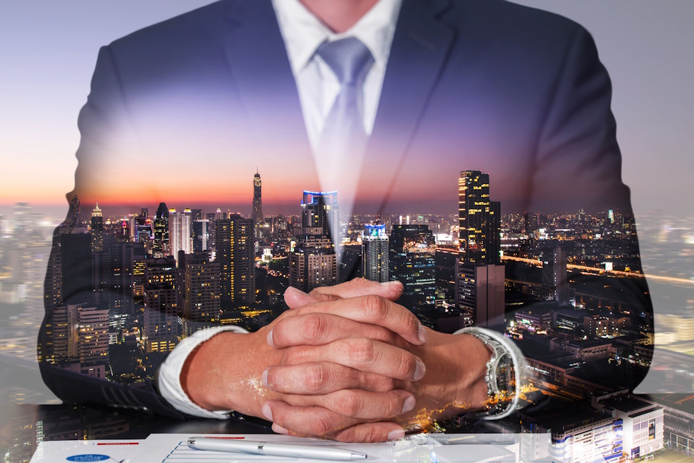 Double exposure of businessman with fingers interlaced and sitting down with a city in the background.